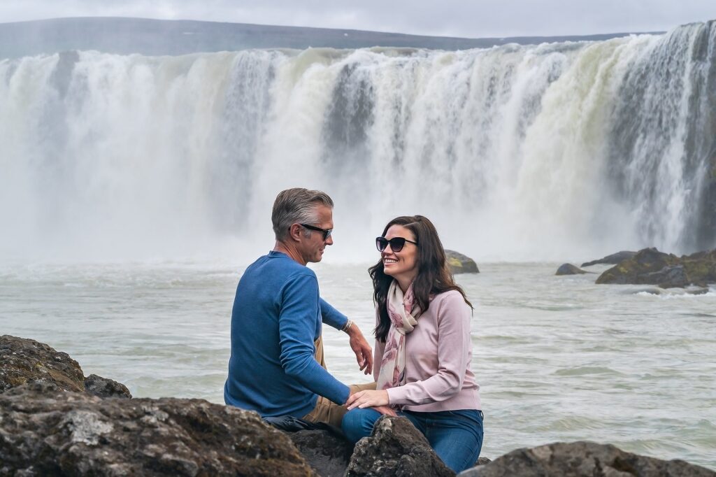 Couple wearing sunglasses while sitting near Godafoss waterfall