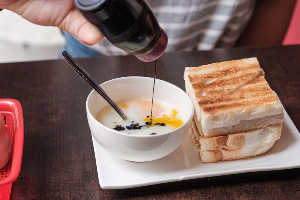 Person adding soy sauce to soft boiled egg with kaya toast