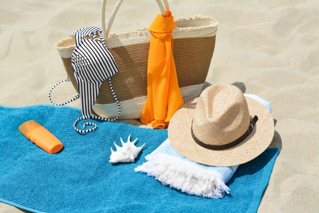 Collection of beach essentials including swimwear, sunscreen, towel, and hat on sand