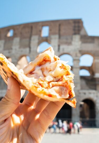 Hand holding a slice of Italian pizza with the Colosseum in the background