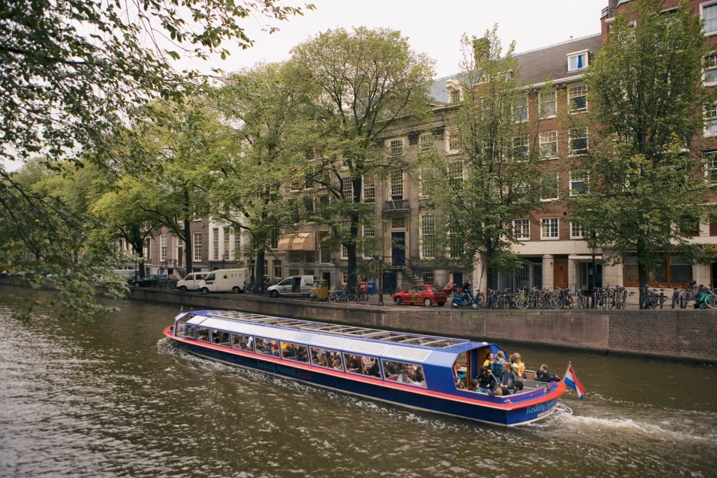 Glass-roofed tour boat cruising a wide Amsterdam canal facing historic brick houses and numerous bicycles. 