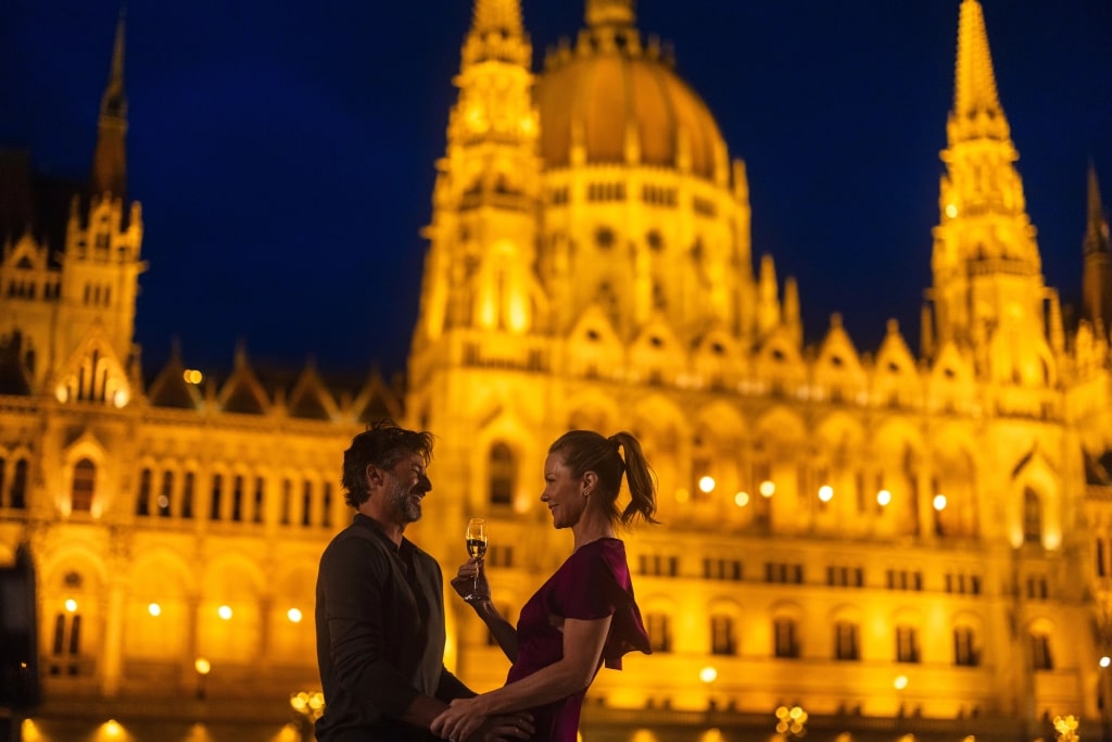A smiling couple poses together, foregrounding the magnificent neo-Gothic Budapest Parliament Building