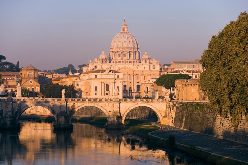 Sunlit Tiber River reflects the stone bridge arches and the iconic, distant towering dome of St. Peter’s Basilica.