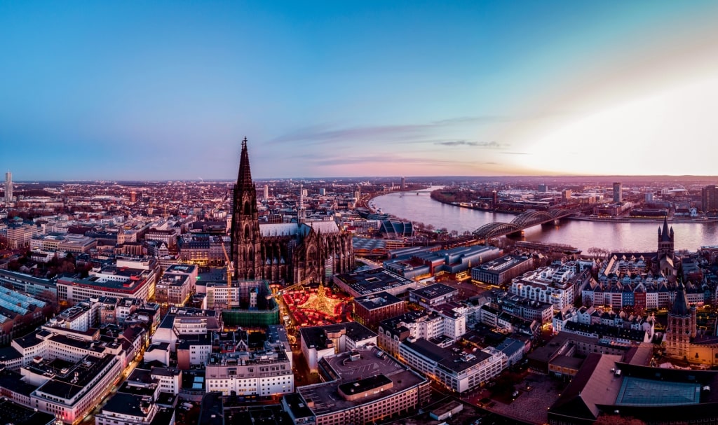 Cologne Cathedral's twin ornate spires and glowing Christmas market dominating the twilight Rhine riverfront.