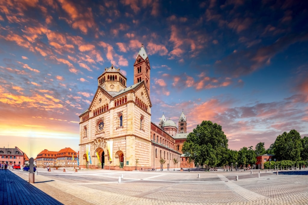 Massive Romanesque Speyer Cathedral dominating a vast stone square facing a colorful sunset sky. 