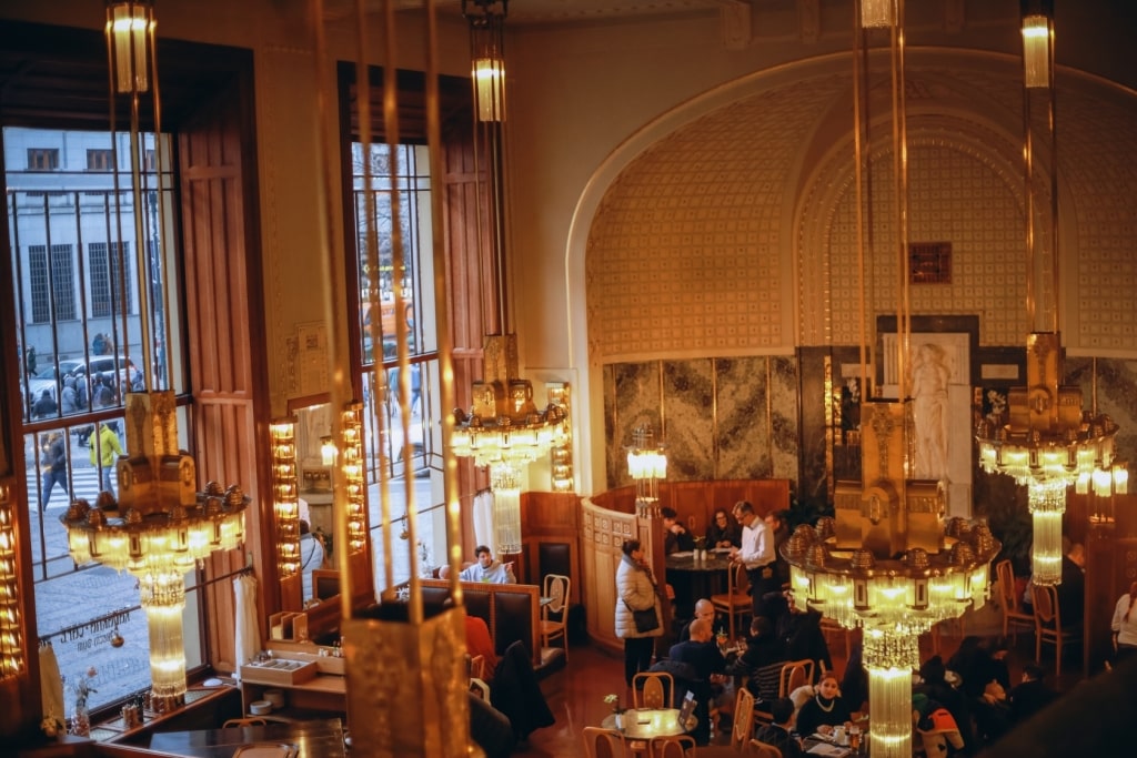 Guests relax beneath ornate Art Deco chandeliers inside a historic café in Prague, Czech Republic.