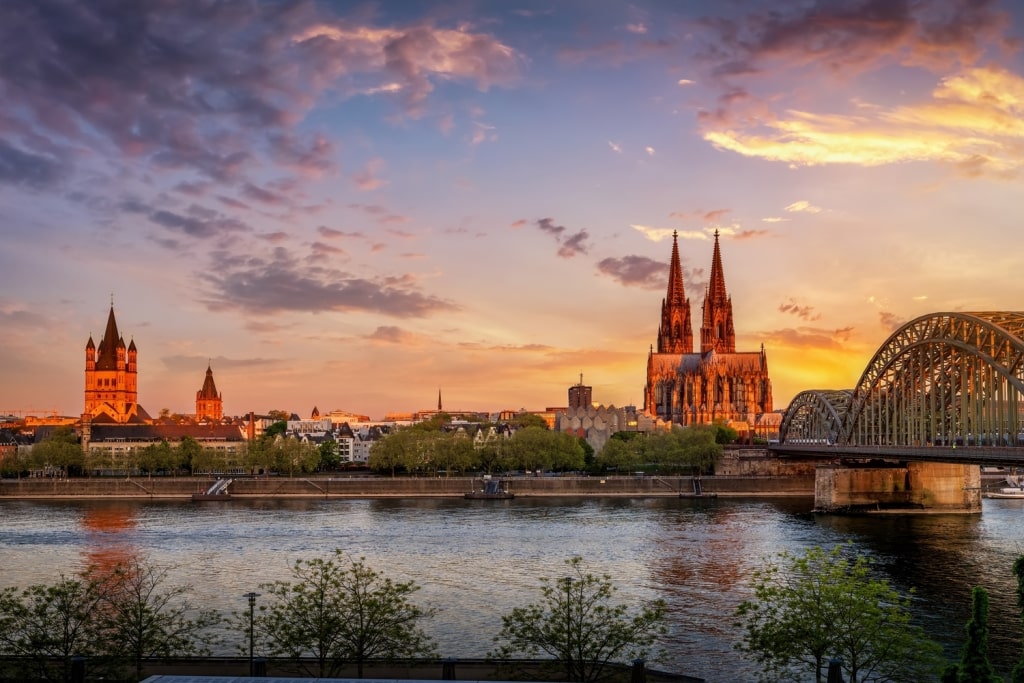 Love-padlock-covered Hohenzollern Bridge framing massive Cologne Cathedral twin spires facing sunset skies.