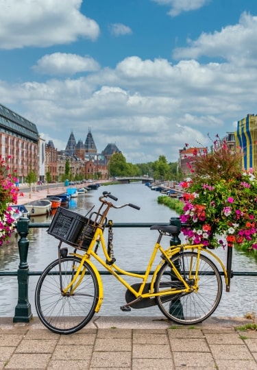 A yellow bicycle parked beside a canal in Amsterdam, Netherlands, framed by vibrant pink flower boxes, with historic canal houses, boats, and a blue sky filled with scattered clouds in the background.