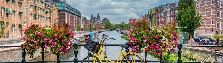 A yellow bicycle parked beside a canal in Amsterdam, Netherlands, framed by vibrant pink flower boxes, with historic canal houses, boats, and a blue sky filled with scattered clouds in the background.
