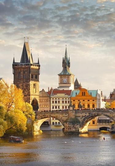 Golden sunrise over Charles Bridge spanning the Vltava River in Prague, Czech Republic, with the Old Town skyline, historic towers, colorful buildings, and autumn trees reflected in the water.