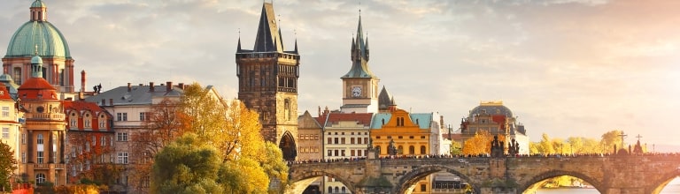 Golden sunrise over Charles Bridge spanning the Vltava River in Prague, Czech Republic, with the Old Town skyline, historic towers, colorful buildings, and autumn trees reflected in the water.