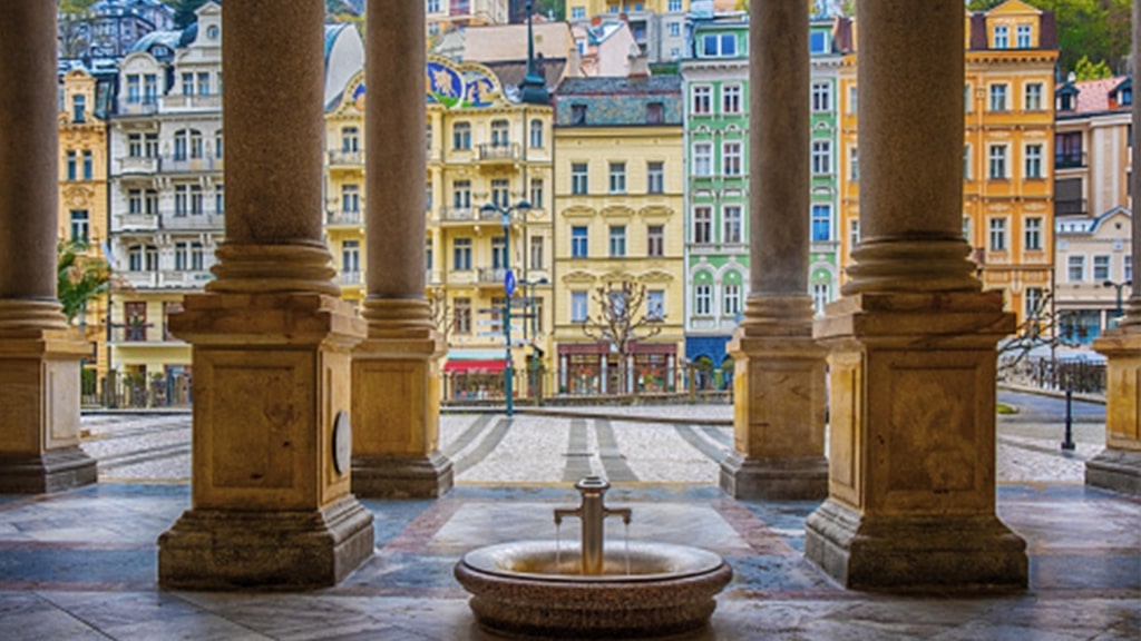 Stone columns frame a mineral water fountain within the colorful Karlovy Vary buildings and beyond.