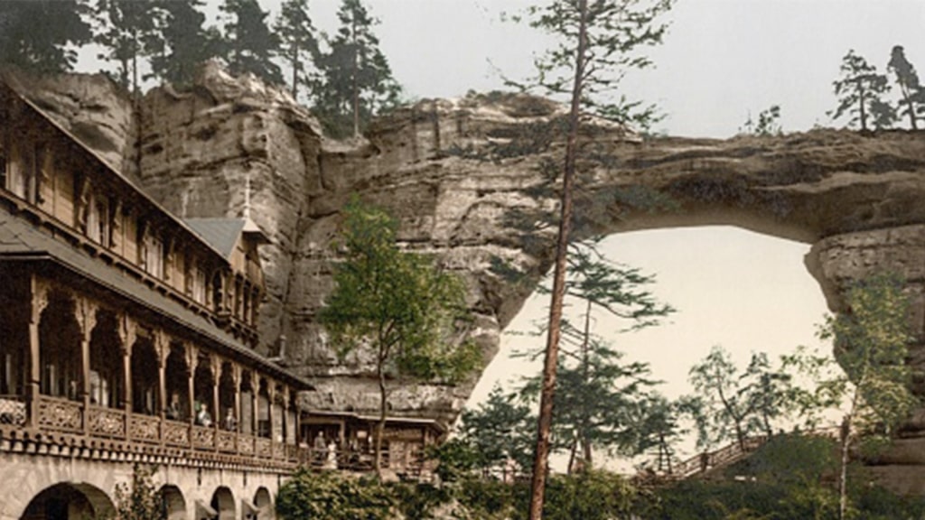 The monumental sandstone arch of Pravčická Gate towers beside a historic wooden lodge.