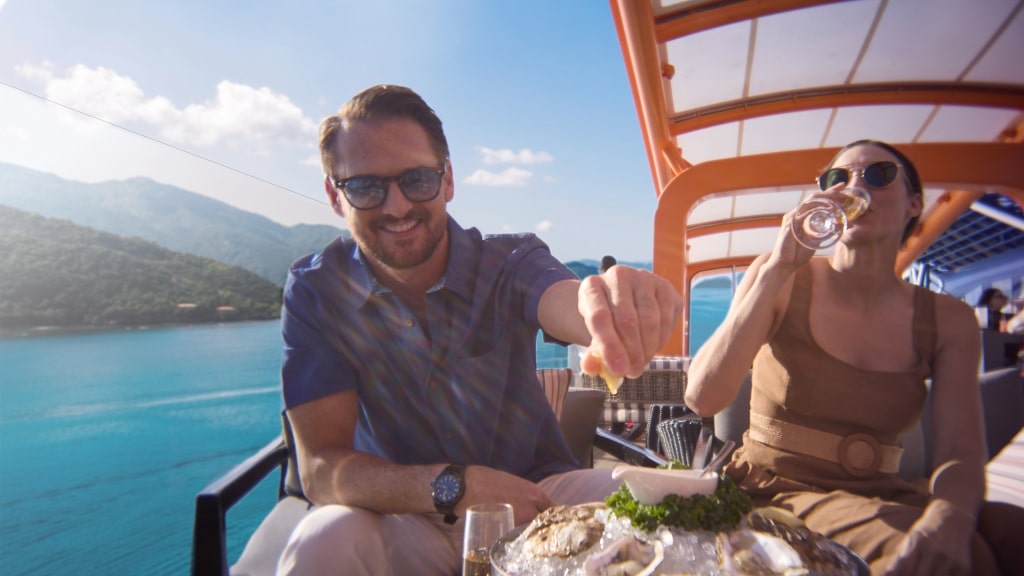 A couple relaxes on an outdoor deck aboard a Celebrity cruise ship, enjoying fresh oysters and sparkling wine while overlooking turquoise waters and lush tropical mountains on a sunny day.