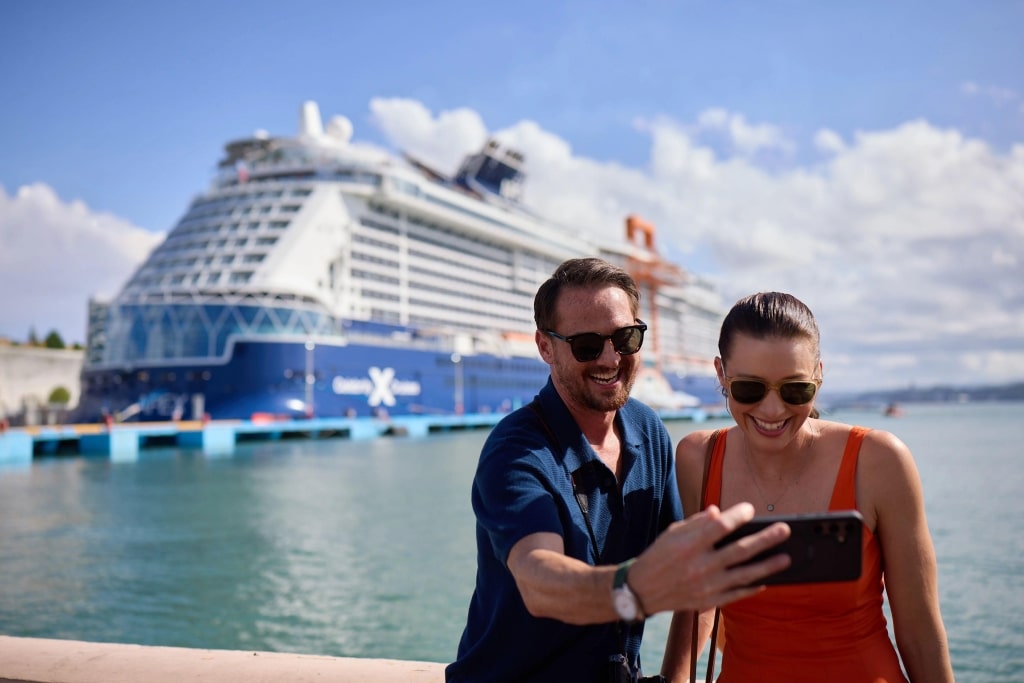 A smiling couple poses in the foreground, overlooking a majestic distant Celebrity cruise ship docking.