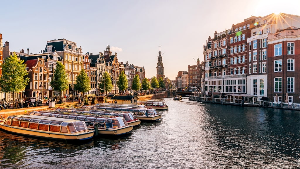 Tour boats line an Amsterdam canal surrounded by historic buildings under a glowing sunburst.