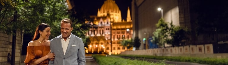 A couple shares a warm smile while strolling at night before the illuminated dome of the Hungarian Parliament Building.