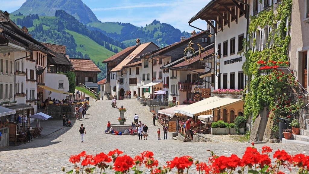 Red flowers frame the cobblestone street of medieval Gruyères beneath lush green mountains.