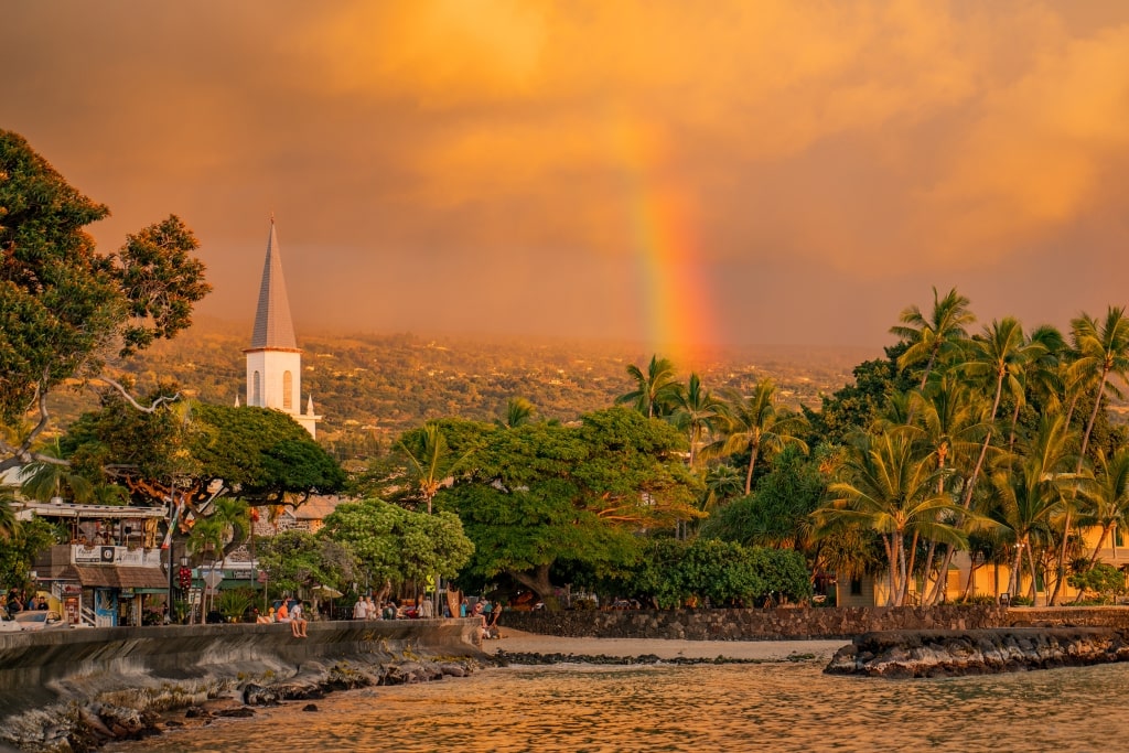 A vibrant rainbow arches over lush palm trees and the iconic church spire of Kailua-Kona at sunset.