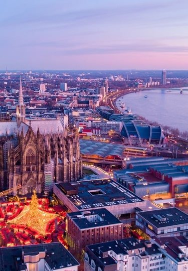 An aerial view reveals the glowing Christmas market sprawling at the foot of Cologne Cathedral along the twilight Rhine River.