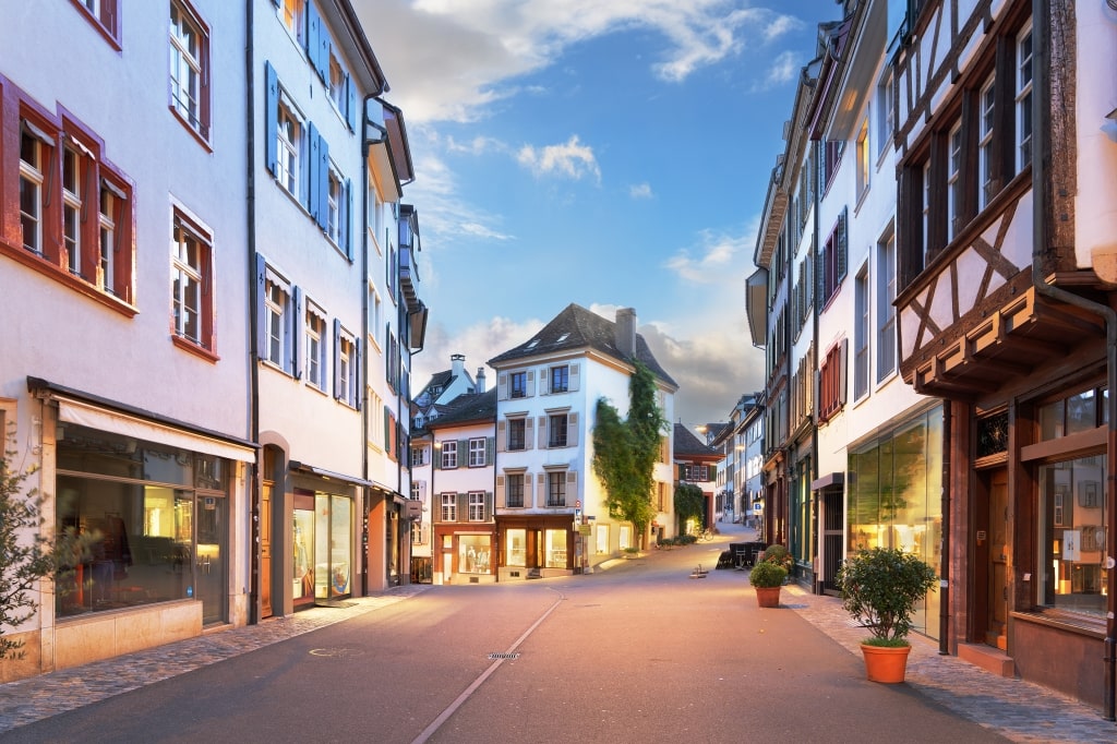 Warmly lit storefronts and historic half-timbered buildings line a quiet street in Basel's Old Town at twilight.