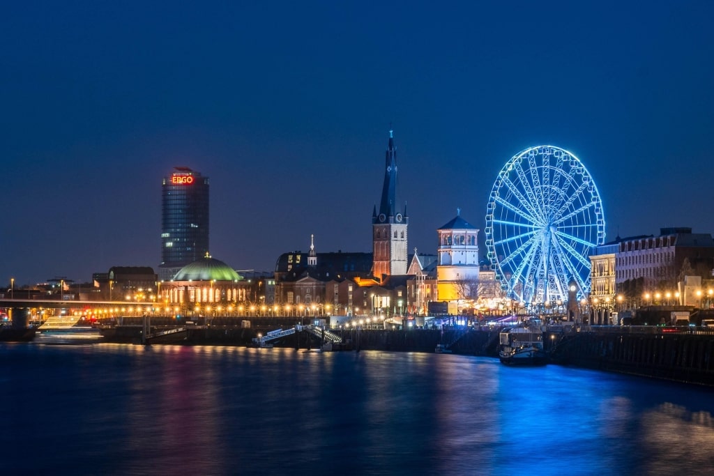 An illuminated Rhine promenade at night featuring the ERGO Tower, a blue illuminated Ferris wheel, and historic landmarks