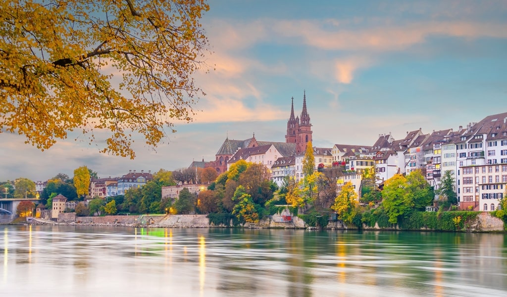 Golden autumn leaves frame the twin spires of the Basel Minster overlooking the calm Rhine River.