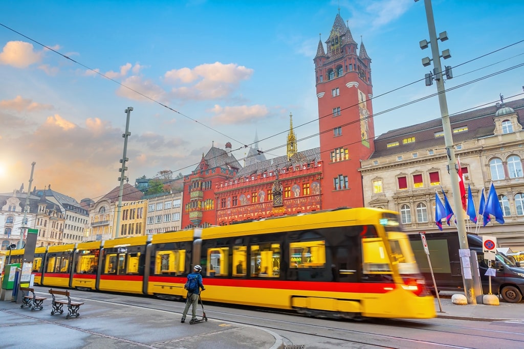 A yellow tram glides past the ornate red facade of the historic Basel Town Hall.