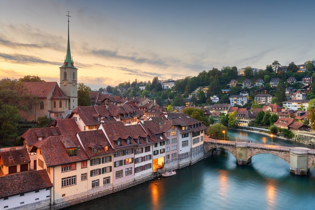 Warm lights illuminate the historic red-roofed buildings and stone bridge along the calm Aare River in Bern at twilight.