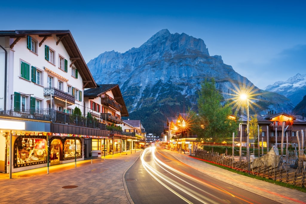 Light trails trace the curving street of Grindelwald past glowing chalets beneath towering Alpine peaks at twilight.