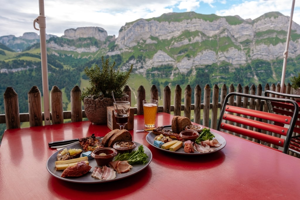 Hearty local platters and drinks sit on a mountain terrace overlooking the dramatic cliffside panorama of Äscher-Ebenalp.