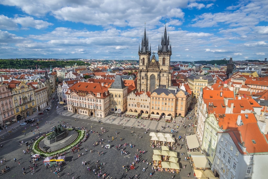 An aerial view captures the twin Gothic spires of the Church of Our Lady before Týn rising above red-tiled roofs and the bustling expanse of Prague's Old Town Square.