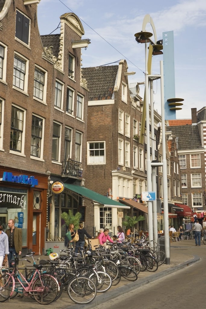 Rows of bikes are stationed along the walkway, set against a backdrop of classic masonry structures and local storefronts in Amsterdam’s historic Nieuwmarkt district.