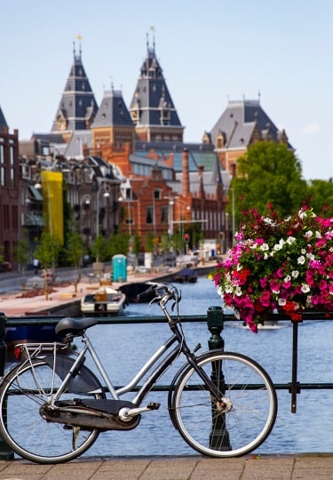A bicycle parked on a canal bridge adorned with vibrant flowers overlooks historic buildings along a waterway in Amsterdam.