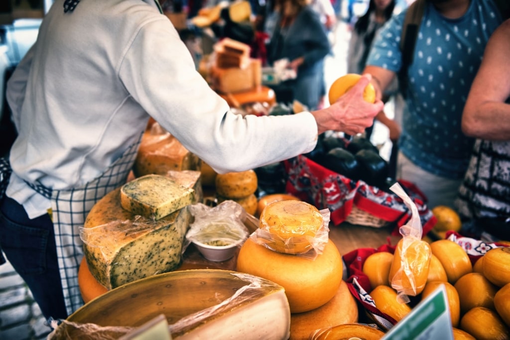 A local merchant offers a petite wheel of artisanal cheese to a visitor amid a vibrant stall showcasing a diverse collection of historic Dutch varieties.