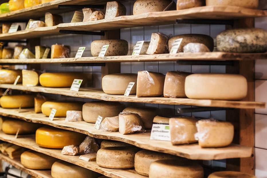A wide variety of traditional Dutch cheese wheels and wedges are displayed across wooden shelves inside a cheese market in Amsterdam.