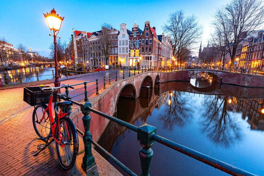 A red bicycle leans against the railing of a bridge, offering a view of serene canal reflections and historic canal houses during the Amsterdam blue hour.