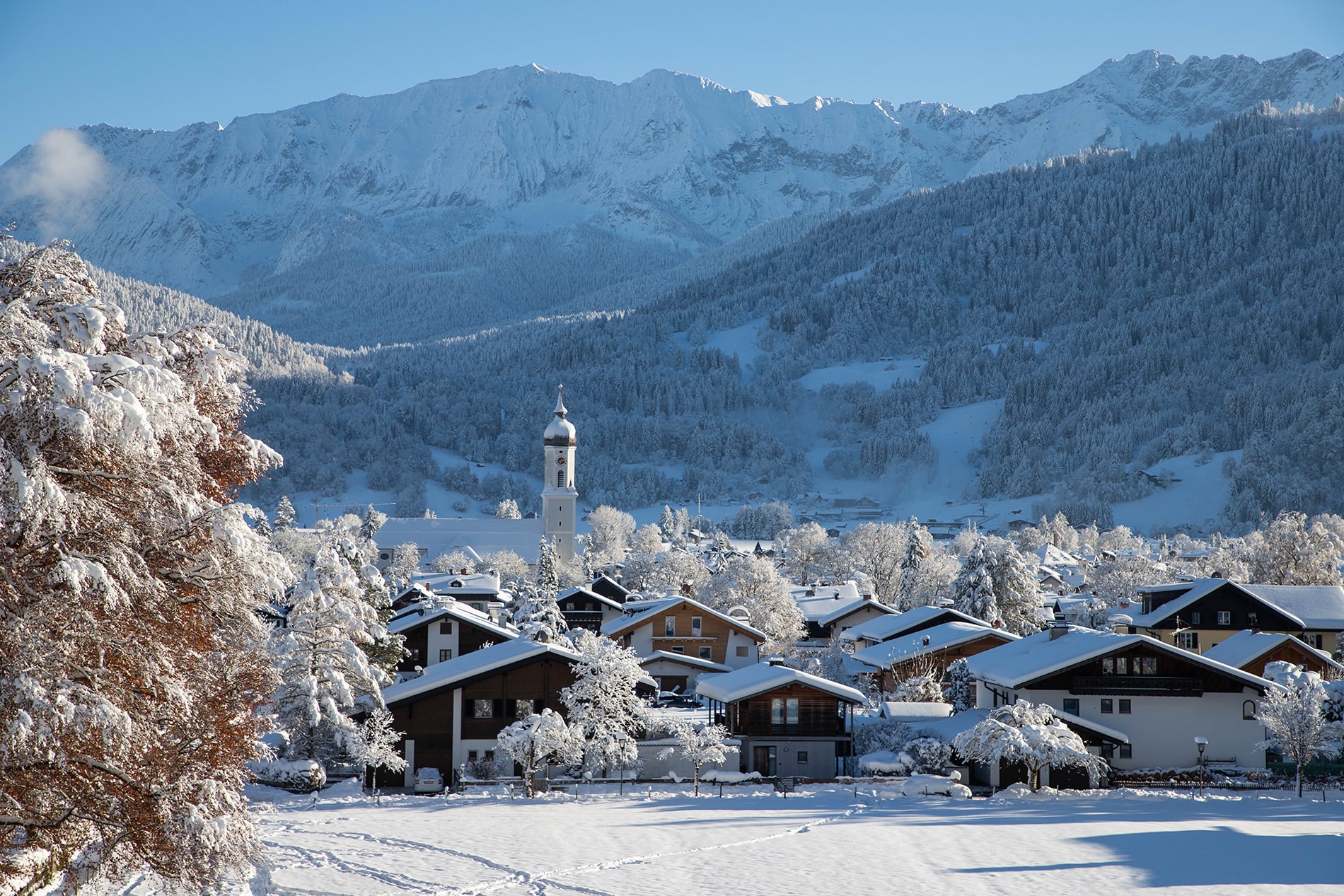 A gorgeous snow-covered village with a distinctive church tower sits nestled beneath the towering, snow-capped peaks of the Bavarian Alps.