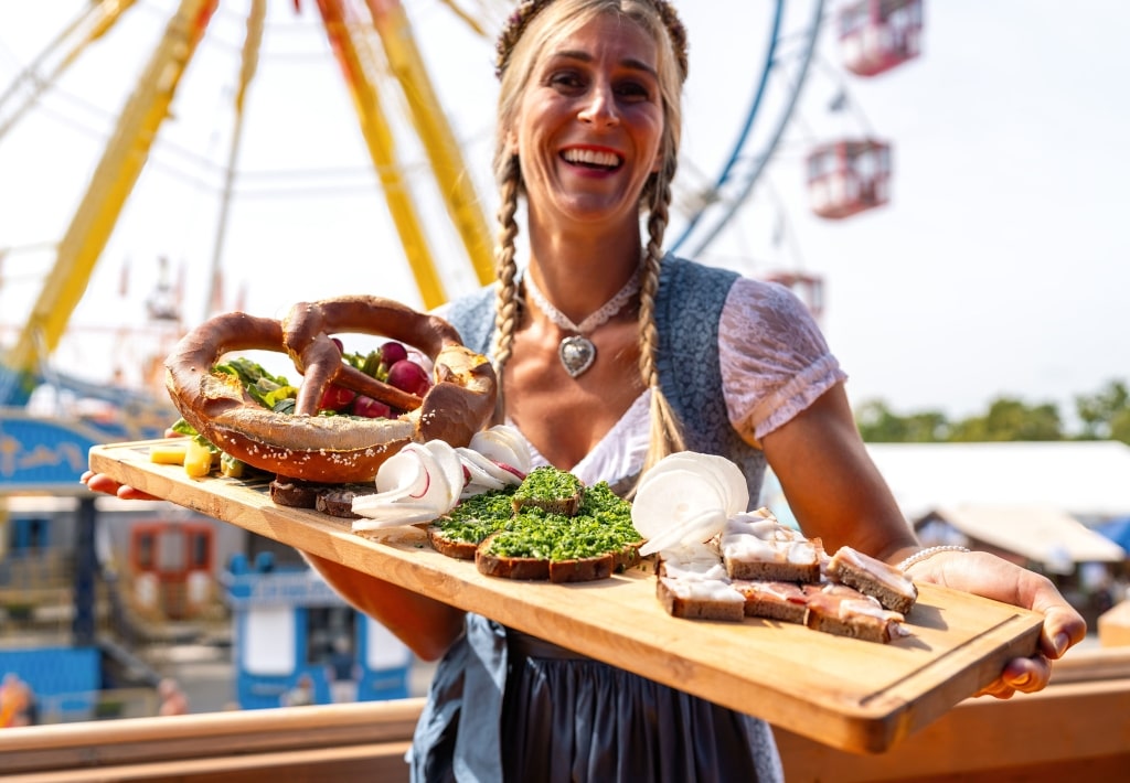 A smiling woman in a dirndl serves German delicacies at an Oktoberfest fairground.