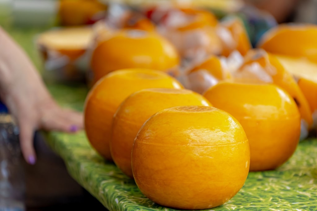 Vibrant spheres of Edam, cloaked in iconic yellow wax, are neatly arranged across a rustic tabletop within a bustling local marketplace.