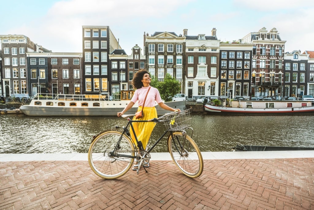 A smiling woman poses with her bicycle along a canal framed by traditional Dutch houses and houseboats in Amsterdam.