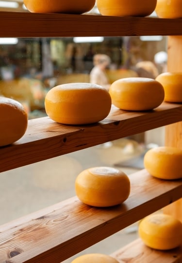 Glistening wheels of Gouda, encased in traditional yellow wax, rest in neat rows atop rustic wooden shelving within a local storefront display.