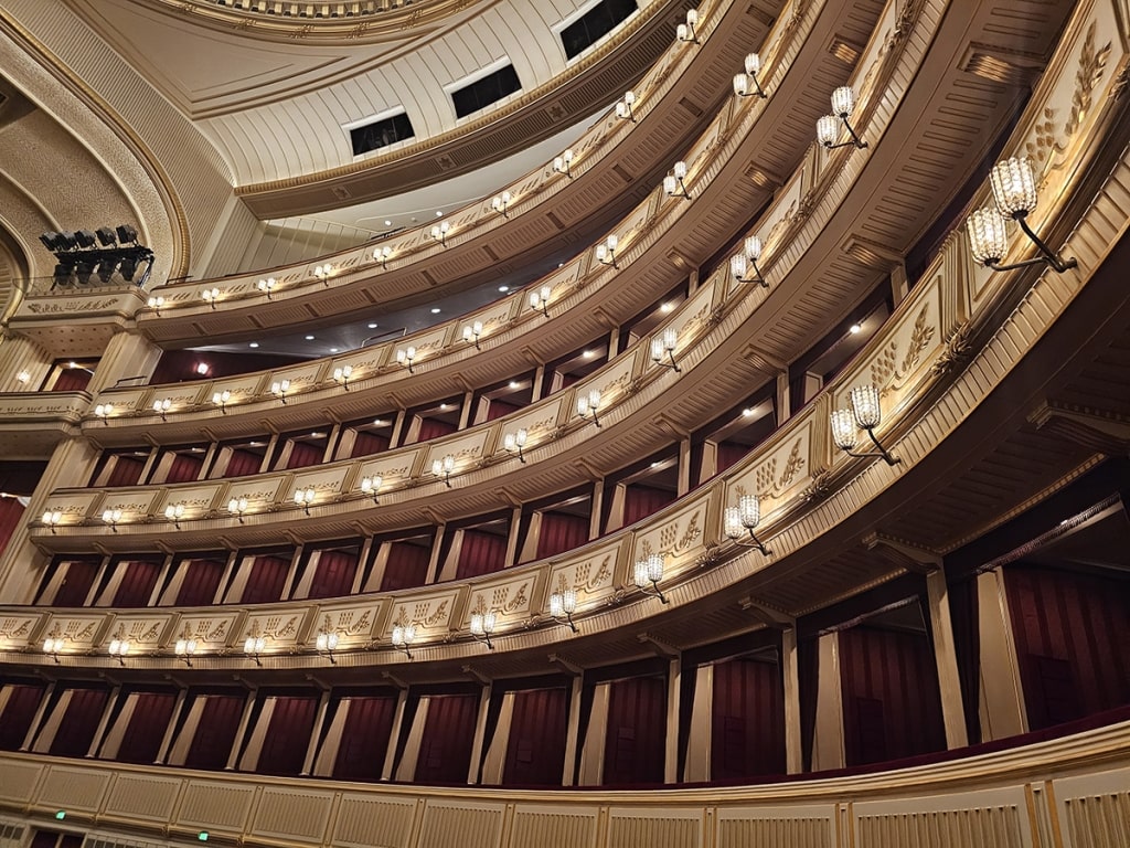 The grand auditorium features glowing, multi-level balconies within the Vienna State Opera.