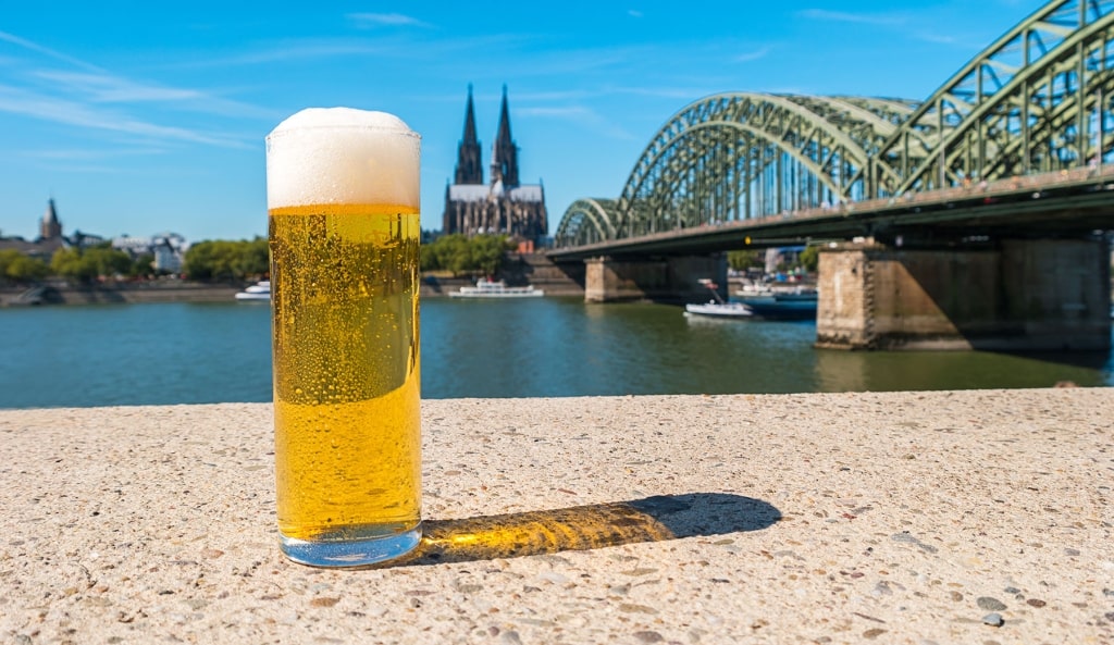 A glass of beer stands before Cologne Cathedral along the Rhine River.