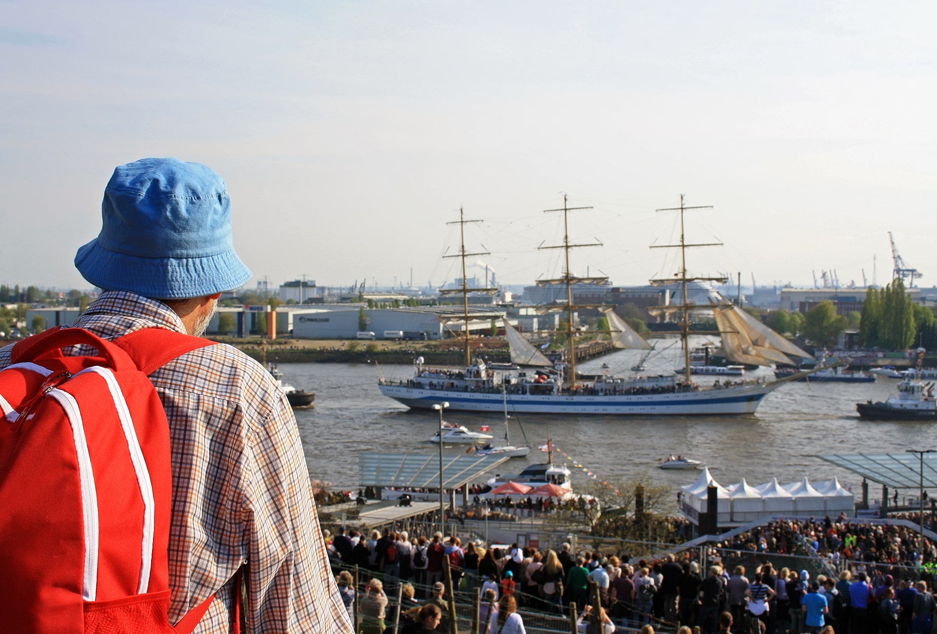 A spectator with a red backpack and blue bucket hat watches a grand tall ship sail down the Elbe River during the Hamburg Port Anniversary.