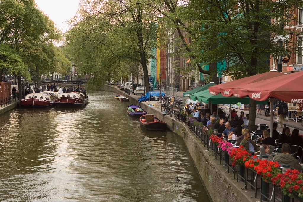 Patrons gather along the flower-lined canal terrace in Amsterdam, capturing the leisurely cadence of water, shade, and sidewalk dining that defines northern European city life.