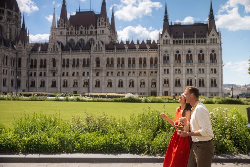 A couple enjoys a sweet chimney cake while strolling past the grand facade of the Hungarian Parliament Building on a sunlit afternoon.