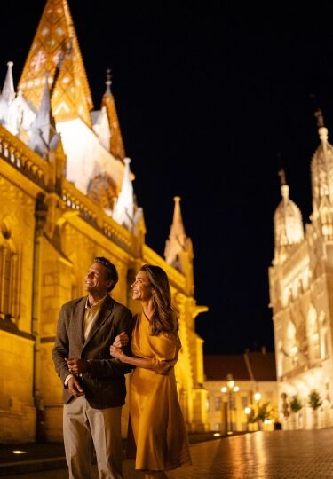 A couple strolls arm-in-arm through Budapest’s Castle District at night, bathed in the warm golden illumination of Matthias Church.
