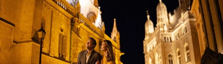 A couple strolls arm-in-arm through Budapest’s Castle District at night, bathed in the warm golden illumination of Matthias Church.