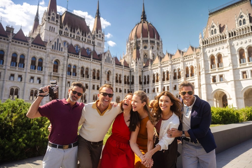 A group of cheerful friends poses together for a photo in front of the grand Hungarian Parliament Building on a sunny summer afternoon.